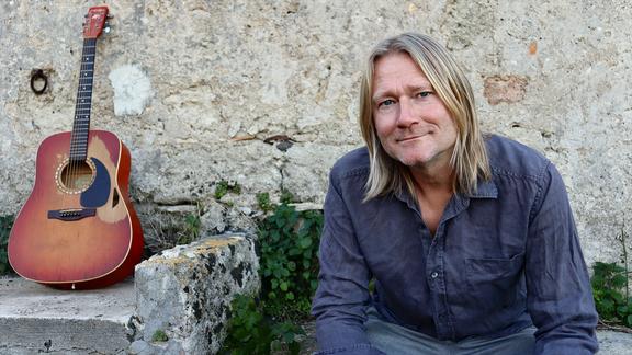 Christian Haase sitzt vor einer Mauer, neben ihm steht seine Gitarre.