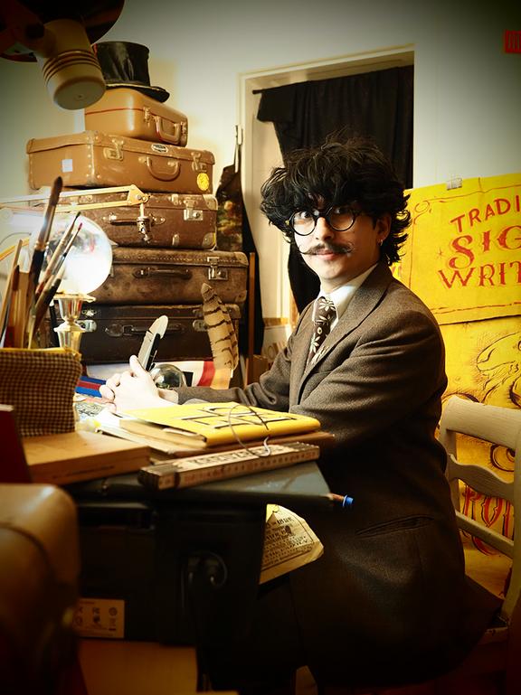 Ernesto Lukas sitzt an einem Schreibtisch umgeben von Requisiten.